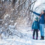 Ein Erwachsener und ein Kind in Winterkleidung gehen auf einem verschneiten Weg durch die Bäume, wobei der Erwachsene auf etwas in den Zweigen zeigt.