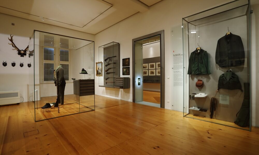 Eine Museumsausstellung zeigt historische Uniformen, Jagdausrüstungen und Geweihe in Glasvitrinen auf polierten Holzböden mit gerahmten Bildern an den Wänden.