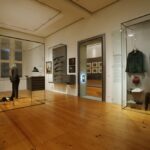 Eine Museumsausstellung zeigt historische Uniformen, Jagdausrüstungen und Geweihe in Glasvitrinen auf polierten Holzböden mit gerahmten Bildern an den Wänden.