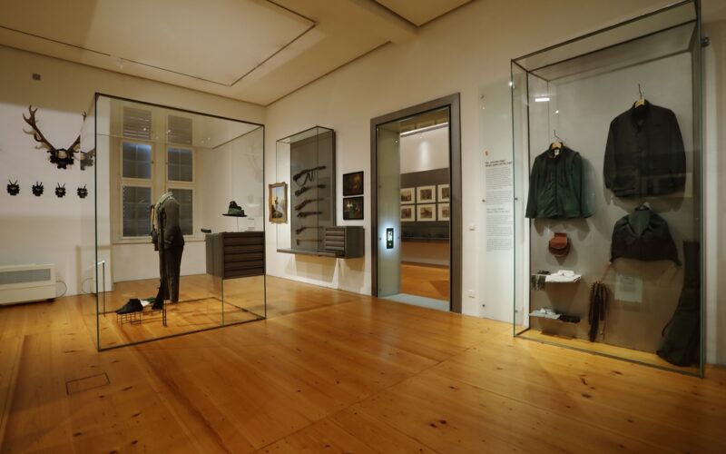 Eine Museumsausstellung zeigt historische Uniformen, Jagdausrüstungen und Geweihe in Glasvitrinen auf polierten Holzböden mit gerahmten Bildern an den Wänden.