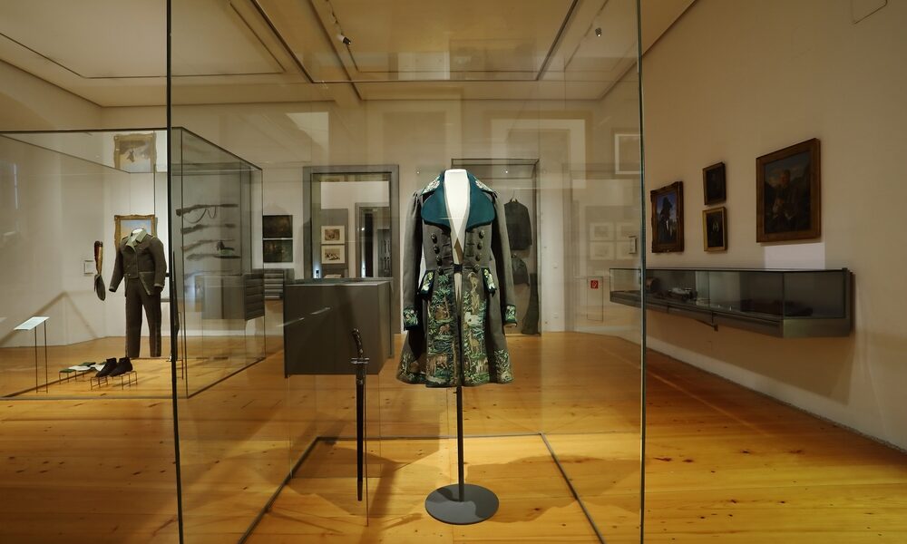 Ein historischer bestickter Mantel, der in einem Museum in einer Glasvitrine ausgestellt ist, mit anderen historischen Kleidungsstücken und Kunstwerken im Hintergrund.