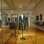 Ein historischer bestickter Mantel, der in einem Museum in einer Glasvitrine ausgestellt ist, mit anderen historischen Kleidungsstücken und Kunstwerken im Hintergrund.