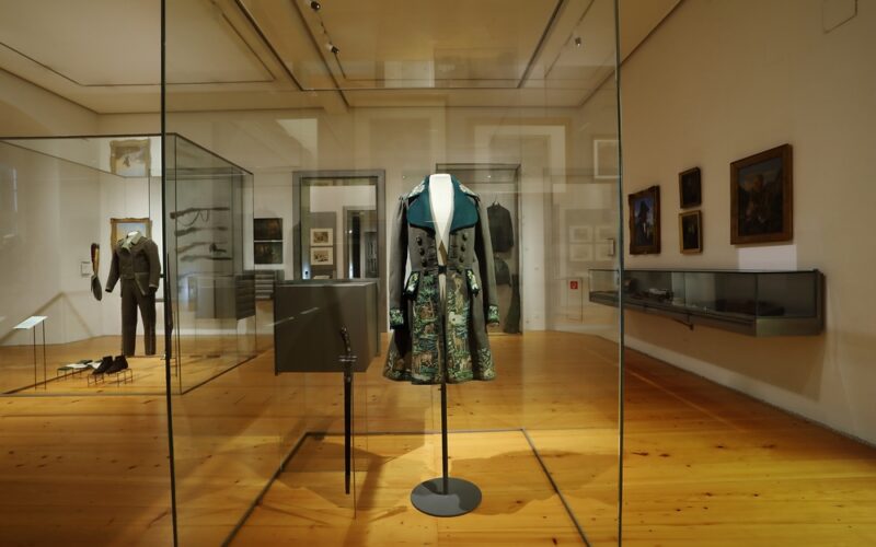 Ein historischer bestickter Mantel, der in einem Museum in einer Glasvitrine ausgestellt ist, mit anderen historischen Kleidungsstücken und Kunstwerken im Hintergrund.