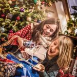 Zwei Kinder sitzen vor einem geschmückten Weihnachtsbaum, lächeln und halten Geschenke, darunter ein Paar Schlittschuhe, umgeben von geöffneten Geschenken und festlichem Geschenkpapier.