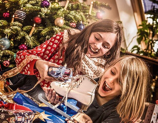 Zwei Kinder sitzen vor einem geschmückten Weihnachtsbaum, lächeln und halten Geschenke, darunter ein Paar Schlittschuhe, umgeben von geöffneten Geschenken und festlichem Geschenkpapier.
