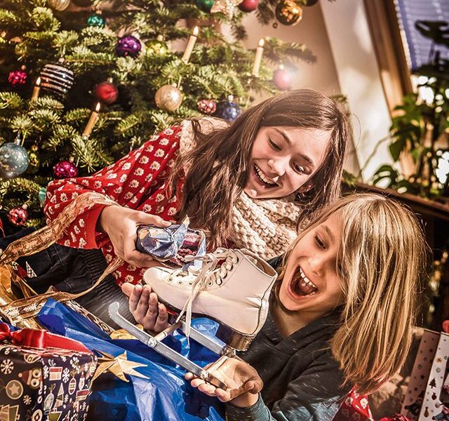 Zwei Kinder sitzen vor einem geschmückten Weihnachtsbaum, lächeln und halten Geschenke, darunter ein Paar Schlittschuhe, umgeben von geöffneten Geschenken und festlichem Geschenkpapier.