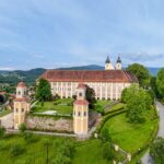 Ein großes historisches Gebäude mit zwei Türmen, umgeben von grünen Bäumen und Gras, befindet sich in der Nähe einer kurvenreichen Straße und einer kleinen Stadt mit Hügeln im Hintergrund.
