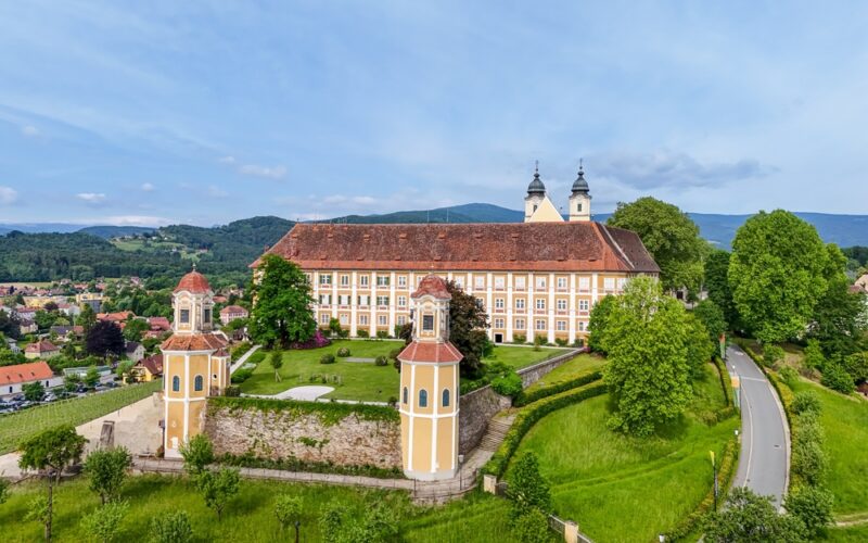Ein großes historisches Gebäude mit zwei Türmen, umgeben von grünen Bäumen und Gras, befindet sich in der Nähe einer kurvenreichen Straße und einer kleinen Stadt mit Hügeln im Hintergrund.