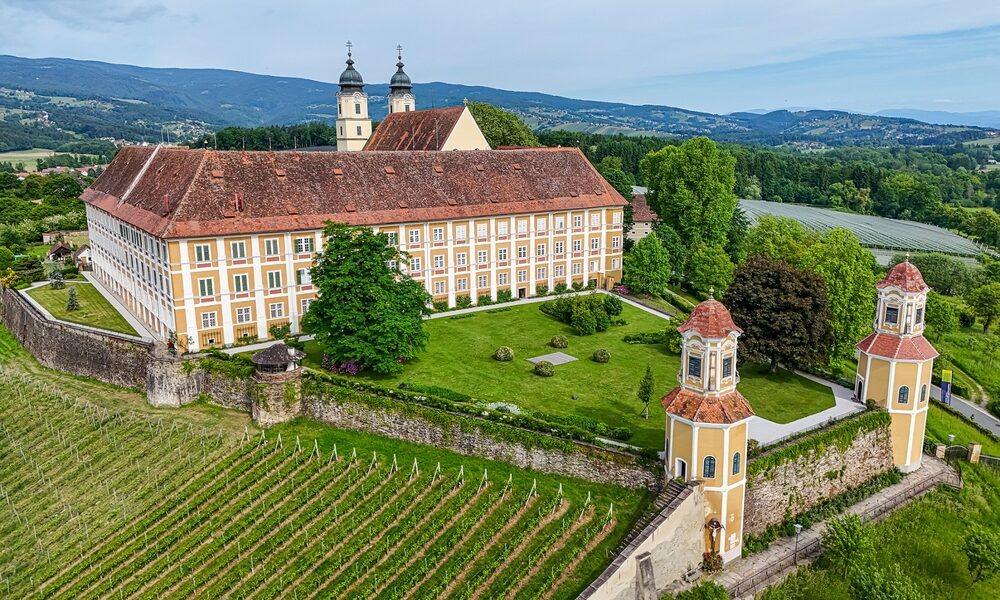 Großes historisches Gebäude mit rotem Ziegeldach, zwei Zwillingstürmen, umgeben von grünen Rasenflächen, Bäumen und Weinbergen, mit Hügeln und bewaldeter Landschaft im Hintergrund.