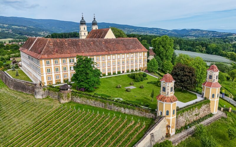 Großes historisches Gebäude mit rotem Ziegeldach, zwei Zwillingstürmen, umgeben von grünen Rasenflächen, Bäumen und Weinbergen, mit Hügeln und bewaldeter Landschaft im Hintergrund.