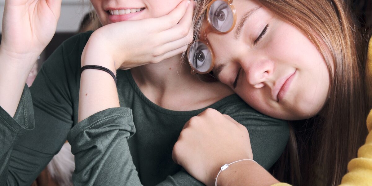 Zwei Mädchen sitzen eng beieinander und halten jeweils eine Brille mit aufgedruckten Augen über ihr Gesicht. Ein Brillenetui mit ähnlichen Brillen liegt auf dem Tisch vor ihnen.