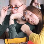 Zwei Mädchen sitzen eng beieinander und halten jeweils eine Brille mit aufgedruckten Augen über ihr Gesicht. Ein Brillenetui mit ähnlichen Brillen liegt auf dem Tisch vor ihnen.