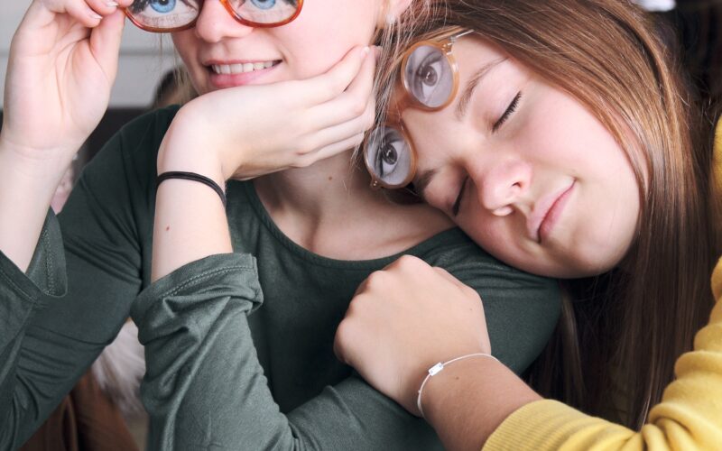 Zwei Mädchen sitzen eng beieinander und halten jeweils eine Brille mit aufgedruckten Augen über ihr Gesicht. Ein Brillenetui mit ähnlichen Brillen liegt auf dem Tisch vor ihnen.