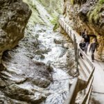 Drei Personen gehen auf einem schmalen Holzsteg entlang eines felsigen Baches in einer Schlucht mit steilen Felswänden und Grünflächen.