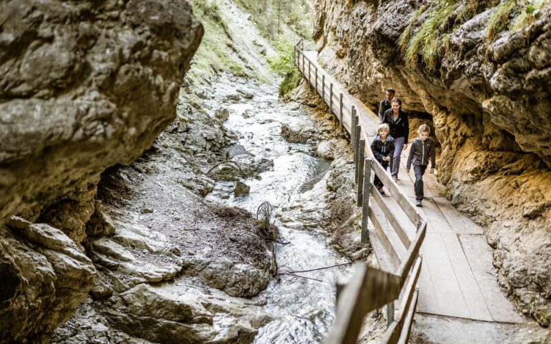 Drei Personen gehen auf einem schmalen Holzsteg entlang eines felsigen Baches in einer Schlucht mit steilen Felswänden und Grünflächen.