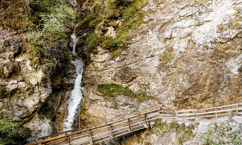 Ein hölzerner Steg überquert einen schmalen Bach unter felsigen Klippen und spärlichem Grün in einer bergigen Gegend.