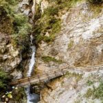 Ein hölzerner Steg überquert einen schmalen Bach unter felsigen Klippen und spärlichem Grün in einer bergigen Gegend.