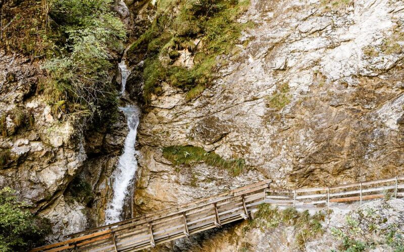 Ein hölzerner Steg überquert einen schmalen Bach unter felsigen Klippen und spärlichem Grün in einer bergigen Gegend.