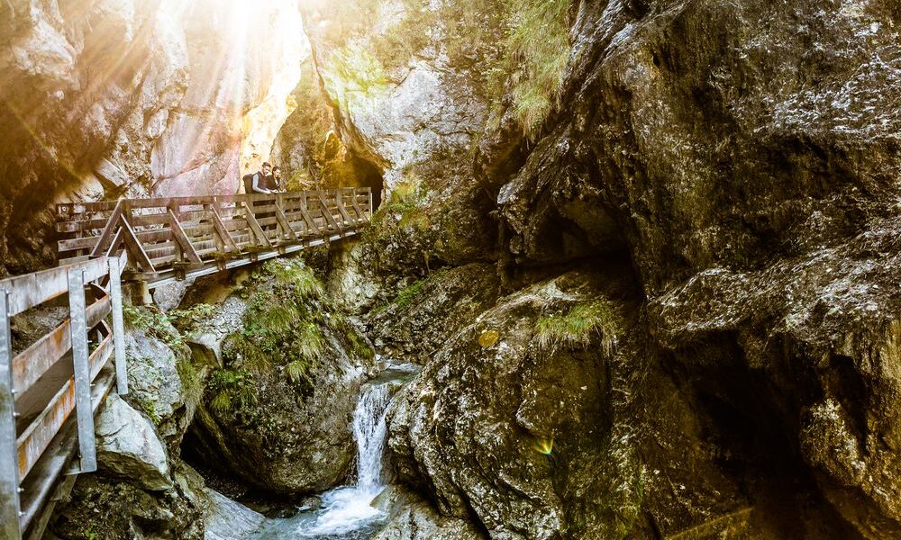 Ein hölzerner Steg überquert eine felsige Schlucht, durch die ein kleiner Bach fließt; das Sonnenlicht fällt durch die Bäume darüber.