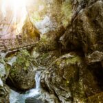 Ein hölzerner Steg überquert eine felsige Schlucht, durch die ein kleiner Bach fließt; das Sonnenlicht fällt durch die Bäume darüber.