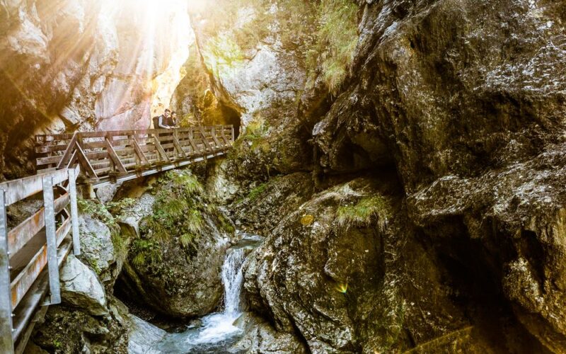 Ein hölzerner Steg überquert eine felsige Schlucht, durch die ein kleiner Bach fließt; das Sonnenlicht fällt durch die Bäume darüber.