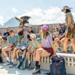 Eine Gruppe von Menschen sitzt auf Bänken im Freien, während ein Hundeführer dem Publikum einen großen Raubvogel vorführt. Im Hintergrund sind Berge und ein Holzzaun zu sehen.