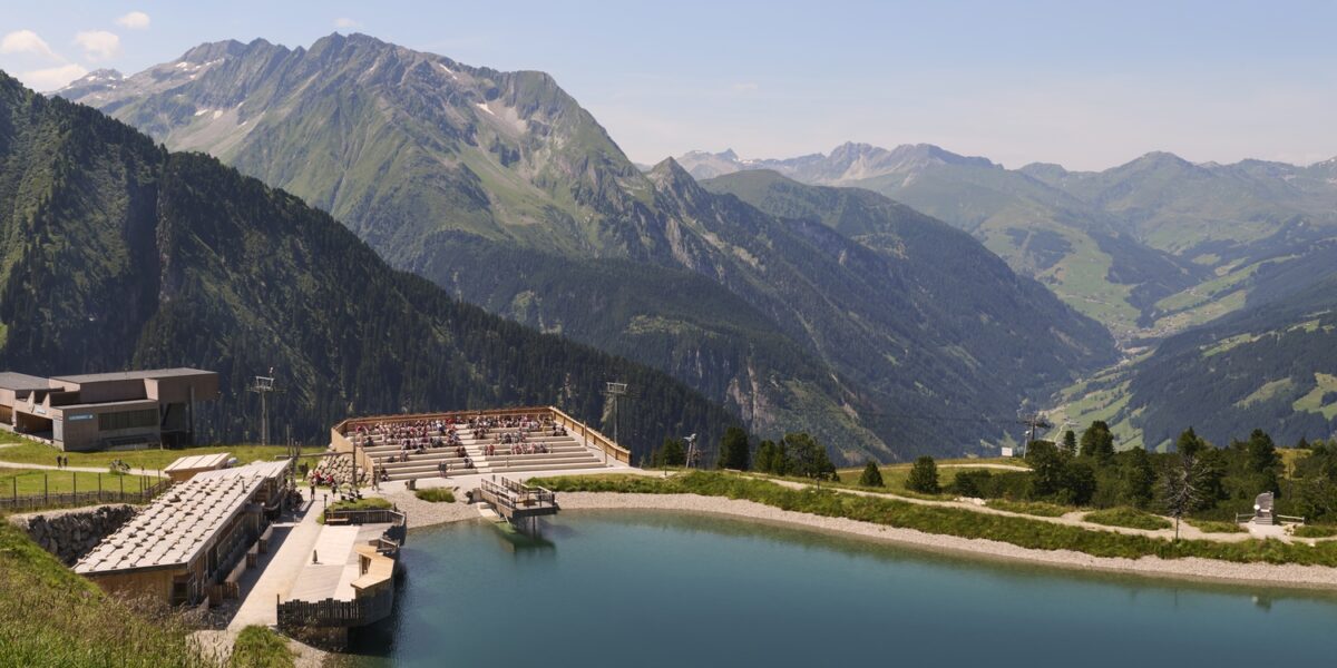 Ein Amphitheater und ein Sitzbereich im Freien bieten einen Blick auf einen ruhigen Bergsee mit grünen Hügeln und fernen Gipfeln unter einem klaren blauen Himmel.