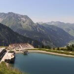 Ein Amphitheater und ein Sitzbereich im Freien bieten einen Blick auf einen ruhigen Bergsee mit grünen Hügeln und fernen Gipfeln unter einem klaren blauen Himmel.