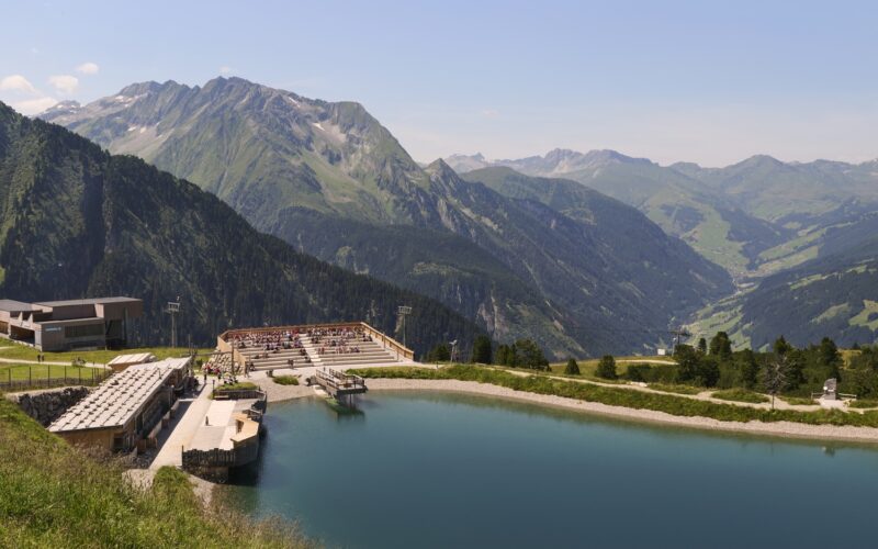 Ein Amphitheater und ein Sitzbereich im Freien bieten einen Blick auf einen ruhigen Bergsee mit grünen Hügeln und fernen Gipfeln unter einem klaren blauen Himmel.