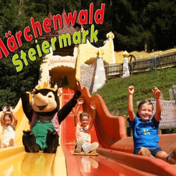 Vier Kinder und eine Maskottchenfigur rutschen im Märchenwald Steiermark, einem Märchenpark, die leuchtend orangefarbenen Rutschen hinunter, mit Bäumen und skurrilen Gebäuden im Hintergrund.