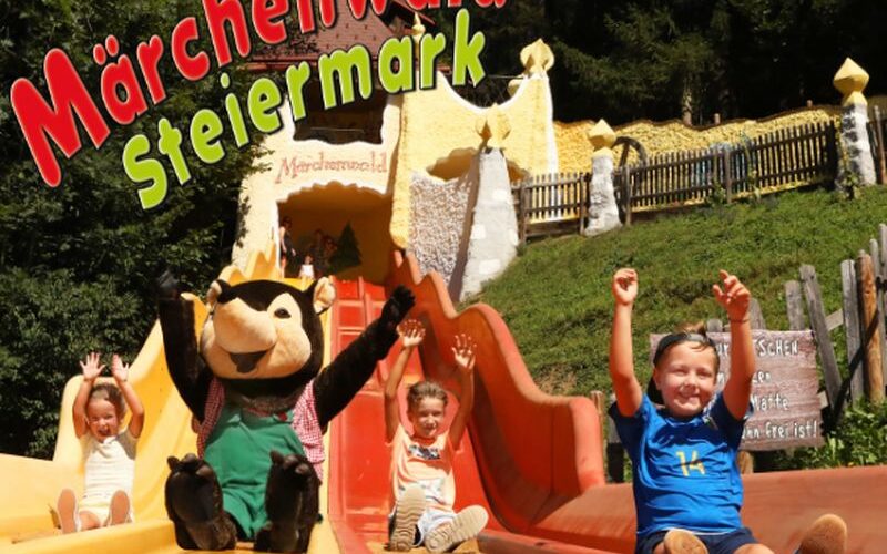 Vier Kinder und eine Maskottchenfigur rutschen im Märchenwald Steiermark, einem Märchenpark, die leuchtend orangefarbenen Rutschen hinunter, mit Bäumen und skurrilen Gebäuden im Hintergrund.