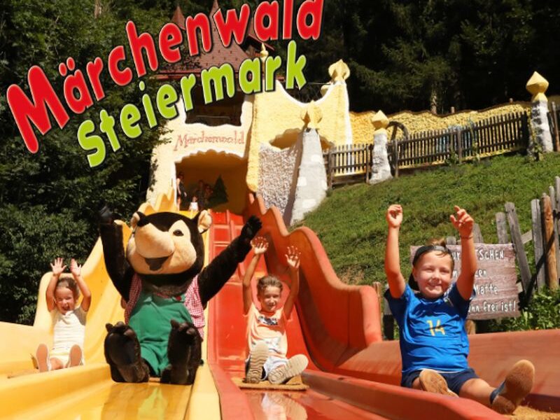 Vier Kinder und eine Maskottchenfigur rutschen im Märchenwald Steiermark, einem Märchenpark, die leuchtend orangefarbenen Rutschen hinunter, mit Bäumen und skurrilen Gebäuden im Hintergrund.