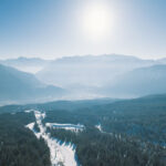 Luftaufnahme eines schneebedeckten Waldes mit Bergen im Hintergrund bei strahlender Sonne und blauem Himmel.