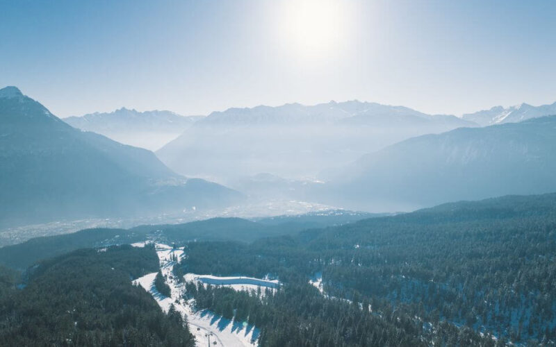 Luftaufnahme eines schneebedeckten Waldes mit Bergen im Hintergrund bei strahlender Sonne und blauem Himmel.