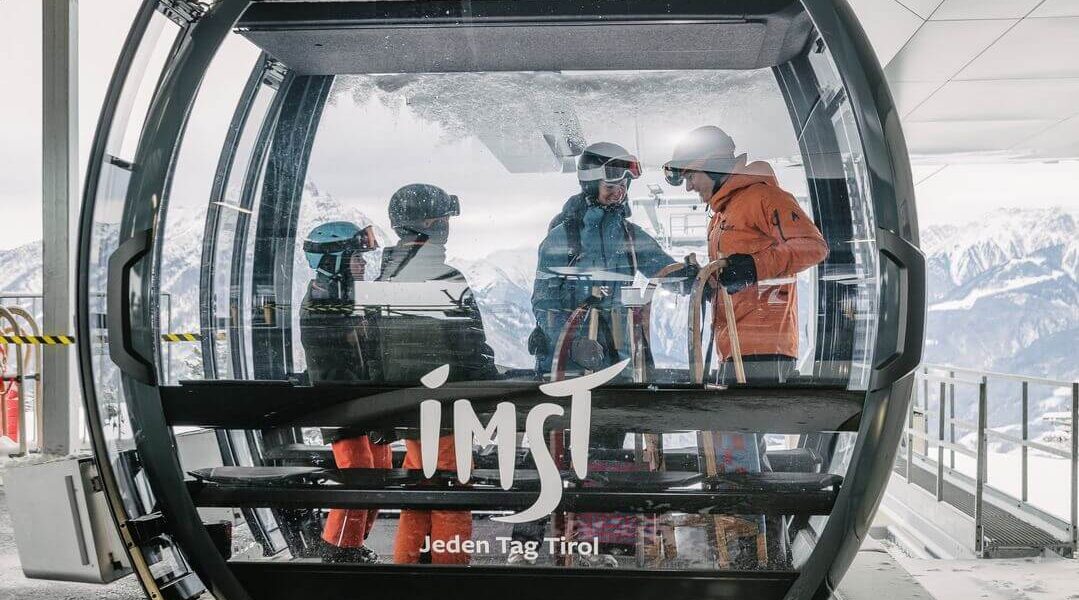 Vier Skifahrer stehen in einer gläsernen Gondel an einer Bergstation eines Skilifts, im Hintergrund sind verschneite Gipfel zu sehen. Die Gondel ist mit "Imst Jeden Tag Tirol" beschriftet.
