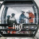 Vier Skifahrer stehen in einer gläsernen Gondel an einer Bergstation eines Skilifts, im Hintergrund sind verschneite Gipfel zu sehen. Die Gondel ist mit "Imst Jeden Tag Tirol" beschriftet.