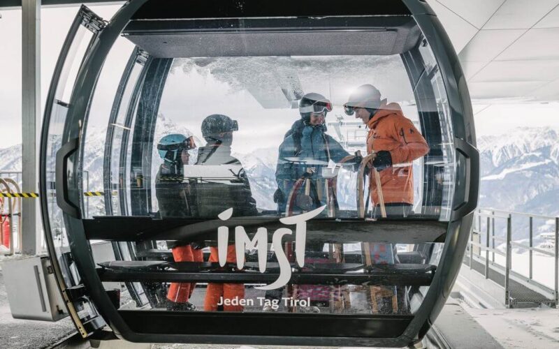 Vier Skifahrer stehen in einer gläsernen Gondel an einer Bergstation eines Skilifts, im Hintergrund sind verschneite Gipfel zu sehen. Die Gondel ist mit "Imst Jeden Tag Tirol" beschriftet.