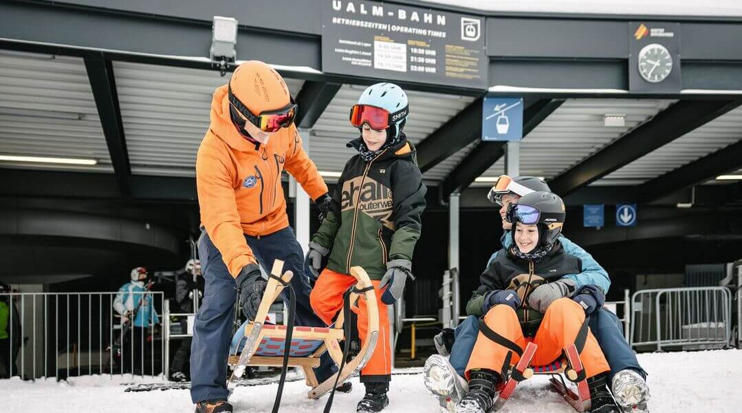 Ein Erwachsener instruiert zwei Kinder auf einem Holzschlitten vor einer Skiliftstation, während ein anderer Erwachsener und ein Kind gemeinsam auf einem roten Schlitten auf verschneitem Boden sitzen.
