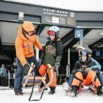 Ein Erwachsener instruiert zwei Kinder auf einem Holzschlitten vor einer Skiliftstation, während ein anderer Erwachsener und ein Kind gemeinsam auf einem roten Schlitten auf verschneitem Boden sitzen.
