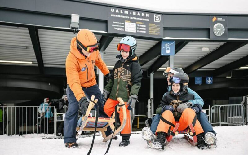 Ein Erwachsener instruiert zwei Kinder auf einem Holzschlitten vor einer Skiliftstation, während ein anderer Erwachsener und ein Kind gemeinsam auf einem roten Schlitten auf verschneitem Boden sitzen.