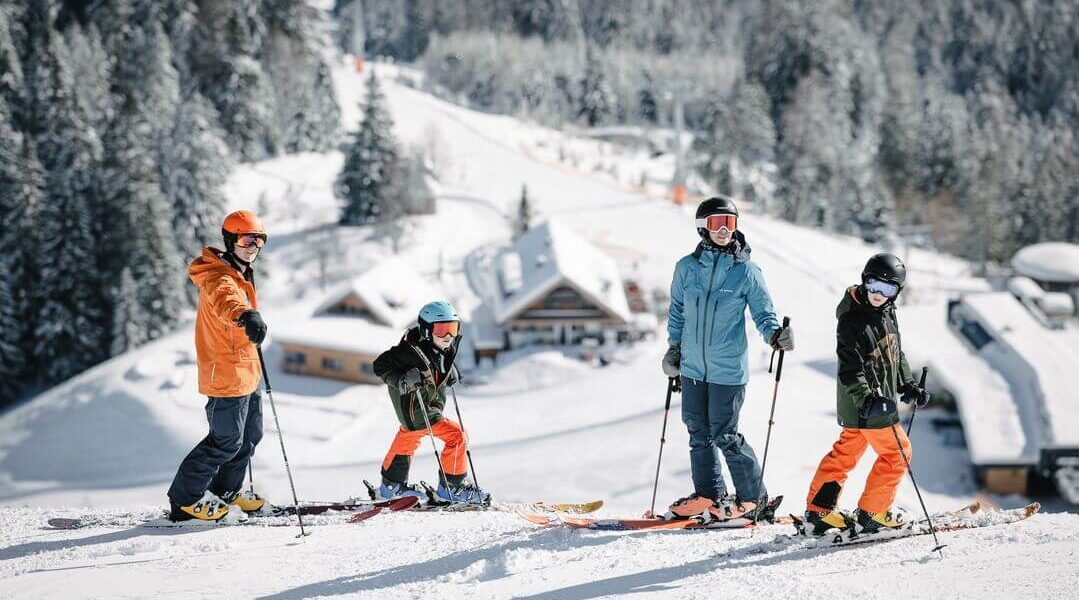Vier Personen in Winterkleidung stehen mit Skiern auf einer verschneiten Piste, im Hintergrund sind Bäume und Gebäude zu sehen.