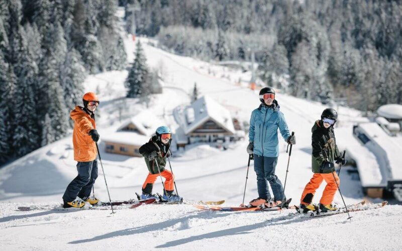 Vier Personen in Winterkleidung stehen mit Skiern auf einer verschneiten Piste, im Hintergrund sind Bäume und Gebäude zu sehen.