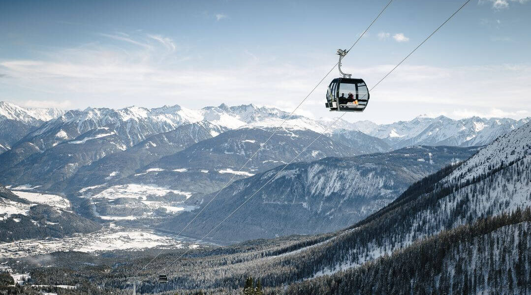 Ein Sessellift fährt über eine schneebedeckte Berglandschaft mit fernen schneebedeckten Gipfeln und einem darunter liegenden Tal hinauf.