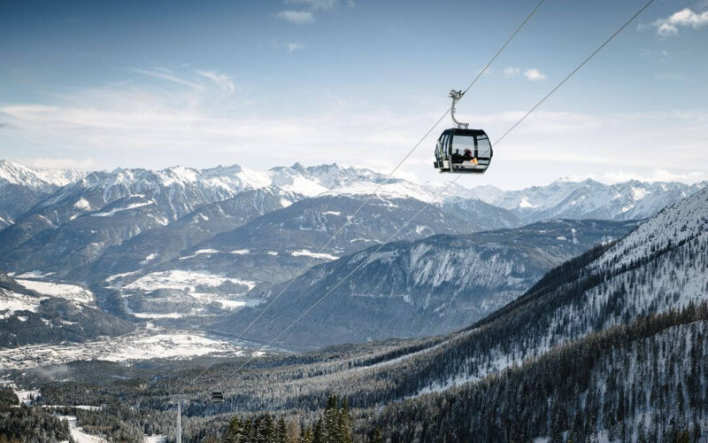 Ein Sessellift fährt über eine schneebedeckte Berglandschaft mit fernen schneebedeckten Gipfeln und einem darunter liegenden Tal hinauf.