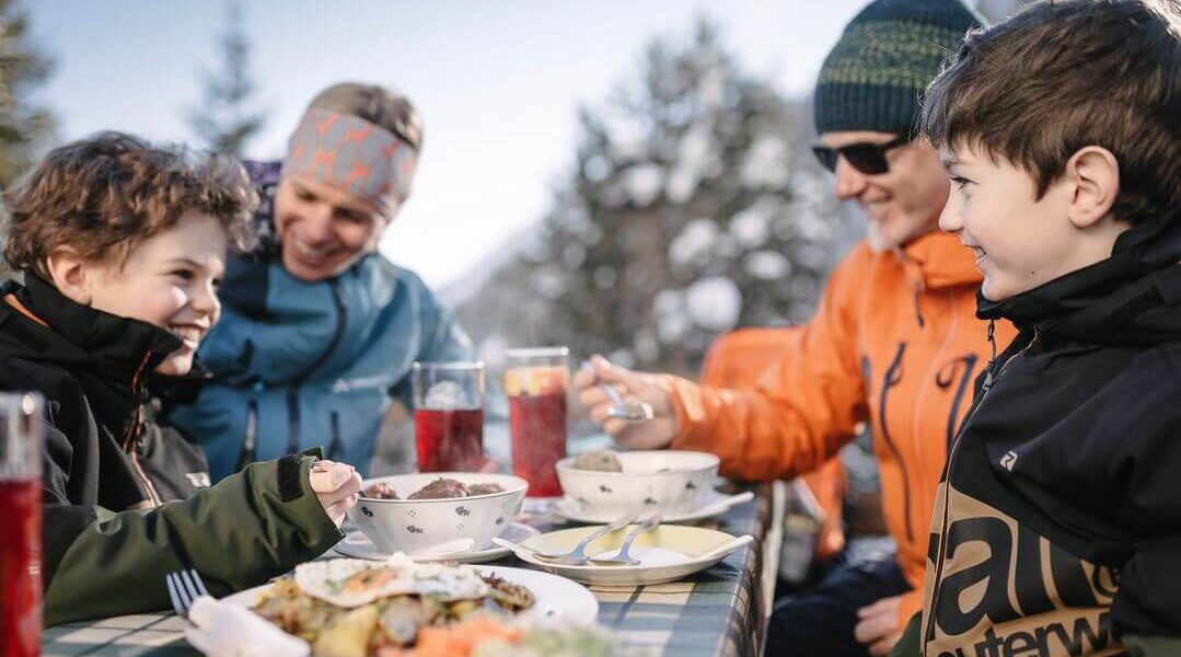 Vier Personen, zwei Erwachsene und zwei Kinder, sitzen an einem sonnigen Tag an einem Tisch im Freien und essen gemeinsam, mit Getränken und Speisen vor sich.