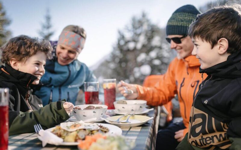 Vier Personen, zwei Erwachsene und zwei Kinder, sitzen an einem sonnigen Tag an einem Tisch im Freien und essen gemeinsam, mit Getränken und Speisen vor sich.