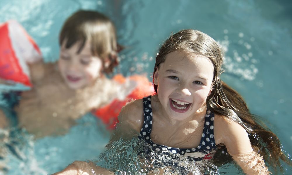Zwei Kinder schwimmen in einem Pool. Das Mädchen im Vordergrund lächelt in die Kamera, während der Junge im Hintergrund orangefarbene Schwimmflügel trägt.
