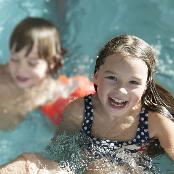 Zwei Kinder schwimmen in einem Pool. Das Mädchen im Vordergrund lächelt in die Kamera, während der Junge im Hintergrund orangefarbene Schwimmflügel trägt.