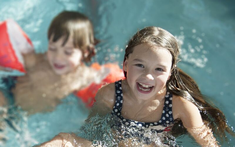 Zwei Kinder schwimmen in einem Pool. Das Mädchen im Vordergrund lächelt in die Kamera, während der Junge im Hintergrund orangefarbene Schwimmflügel trägt.