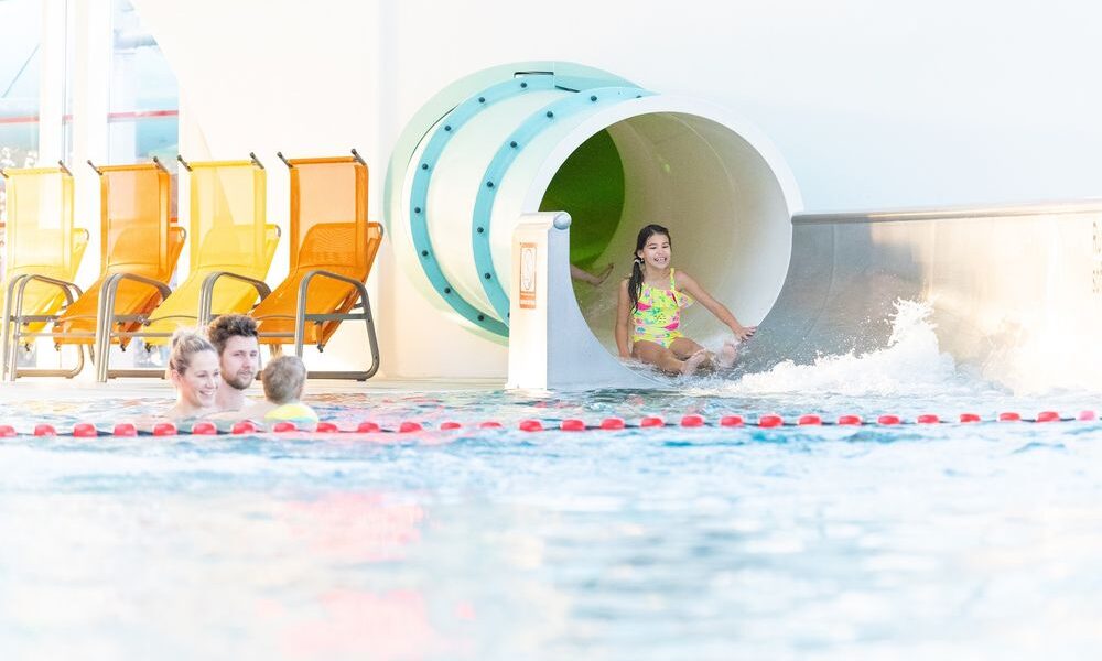 Ein Mädchen steigt von einer Wasserrutsche in ein Schwimmbecken, während drei Personen in der Nähe schwimmen; leere gelbe Stühle säumen den Beckenrand im Hintergrund.